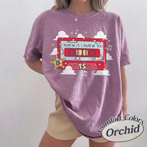 May include: A mauve short-sleeve t-shirt featuring a graphic of a red cassette tape with the text "I KNEW IT, I KNEW YOU" and "TS" inside a yellow star badge. The design includes white clouds, yellow stars, and musical notes surrounding the tape.