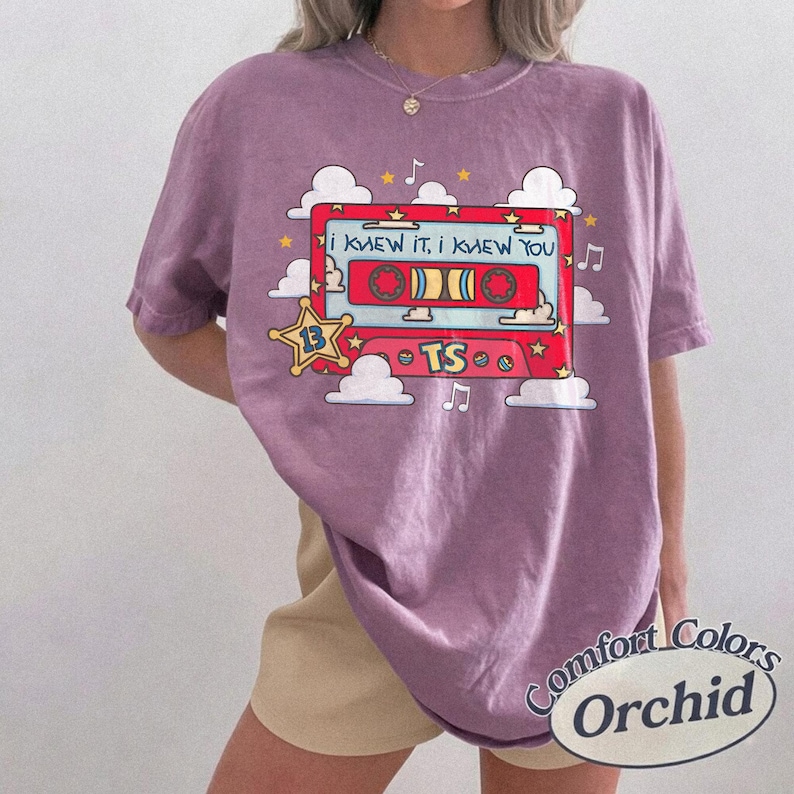 May include: A mauve short-sleeve t-shirt featuring a graphic of a red cassette tape with the text "I KNEW IT, I KNEW YOU" and "TS" inside a yellow star badge. The design includes white clouds, yellow stars, and musical notes surrounding the tape.