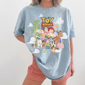 May include: A light blue t-shirt featuring a colorful graphic print of Toy Story characters including Woody, Buzz Lightyear, Jessie, Rex, and others. The design includes white clouds, yellow stars, and the Toy Story logo in red and yellow text.