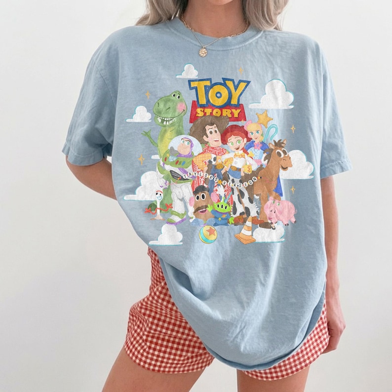 May include: A light blue t-shirt featuring a colorful graphic print of Toy Story characters including Woody, Buzz Lightyear, Jessie, Rex, and others. The design includes white clouds, yellow stars, and the Toy Story logo in red and yellow text.