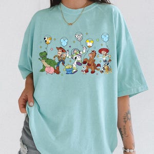 May include: A light blue t-shirt featuring a colorful graphic of animated characters holding balloons. The characters include a green dinosaur, a cowboy, a space ranger, a horse, and a cowgirl. The shirt has short sleeves and a relaxed fit.