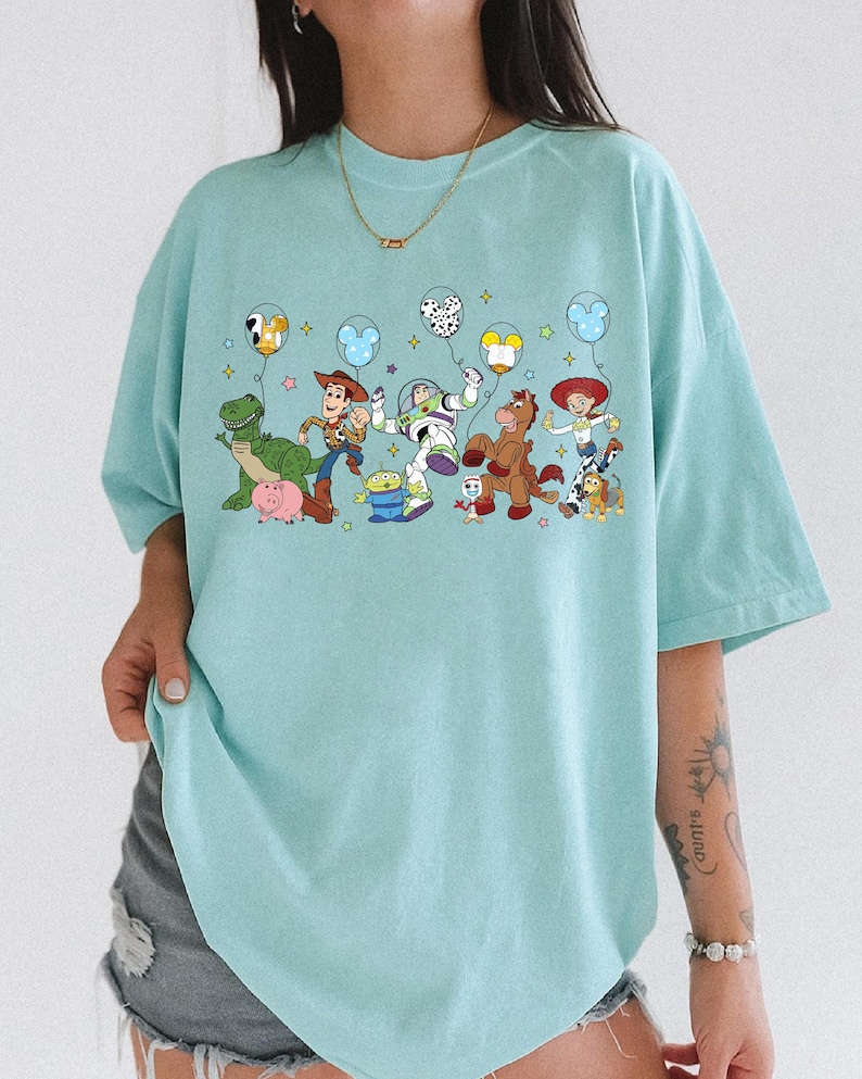 May include: A light blue t-shirt featuring a colorful graphic of animated characters holding balloons. The characters include a green dinosaur, a cowboy, a space ranger, a horse, and a cowgirl. The shirt has short sleeves and a relaxed fit.