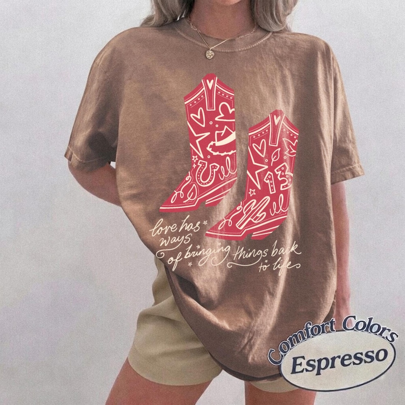 May include: A brown short-sleeve t-shirt featuring a graphic of two red cowboy boots decorated with hearts, stars, and the number 13. Below the boots, the text reads, "love has ways of bringing things back to life" in a cursive white script font.