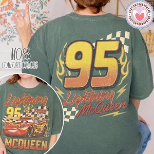 Puede incluir: Camiseta verde con un gran n&uacute;mero 95 en amarillo y naranja, gr&aacute;ficos de llamas y rayos, y el texto "Lightning McQueen". La camiseta tambi&eacute;n tiene el texto "MOSS COMFORT COLORS".