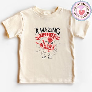 Pode incluir: Camiseta creme com um gr&aacute;fico desgastado do Homem-Aranha. O texto diz "AMAZING SPIDER-MAN" e "Est. '62". O design em vermelho e preto tem um fundo de teia de aranha.