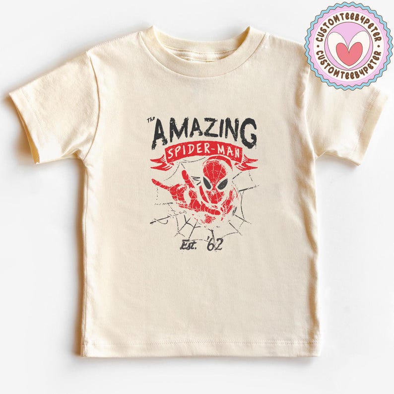 Pode incluir: Camiseta creme com um gr&aacute;fico desgastado do Homem-Aranha. O texto diz "AMAZING SPIDER-MAN" e "Est. '62". O design em vermelho e preto tem um fundo de teia de aranha.