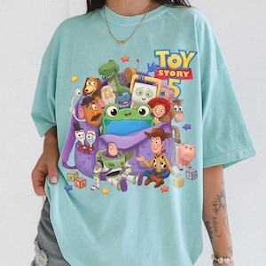 May include: A light blue t-shirt featuring a graphic print of Toy Story 5 characters including Woody, Buzz Lightyear, Jessie, Rex, Slinky Dog, Mr. Potato Head, Forky, and a green frog, all surrounding a purple backpack with heart patterns.
