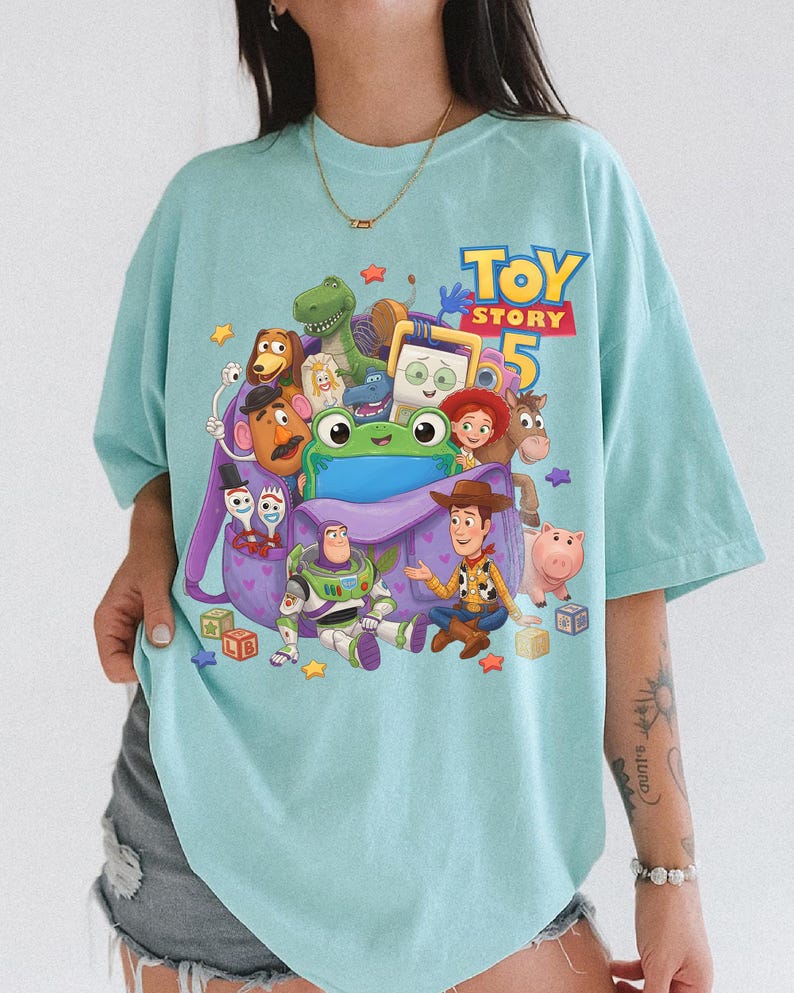 May include: A light blue t-shirt featuring a graphic print of Toy Story 5 characters including Woody, Buzz Lightyear, Jessie, Rex, Slinky Dog, Mr. Potato Head, Forky, and a green frog, all surrounding a purple backpack with heart patterns.