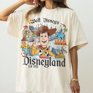 May include: Cream-colored t-shirt featuring a colorful graphic of Woody from Toy Story, Disneyland imagery, and the text "Walt Disney's Disneyland Est 1955." The design includes a train, castle, and other park elements.