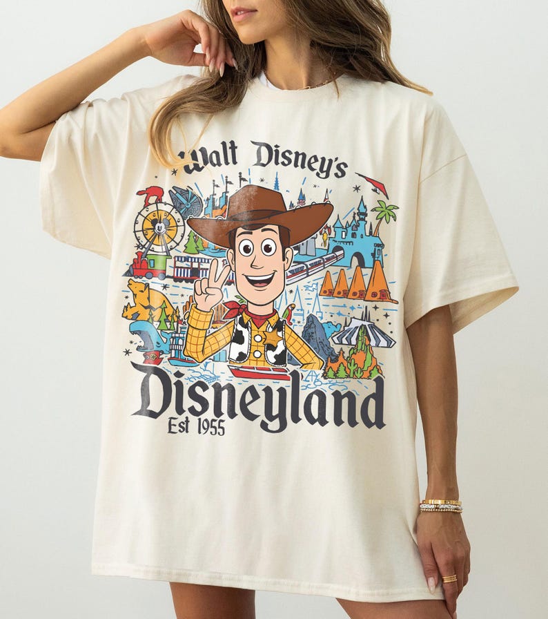 May include: Cream-colored t-shirt featuring a colorful graphic of Woody from Toy Story, Disneyland imagery, and the text "Walt Disney's Disneyland Est 1955." The design includes a train, castle, and other park elements.