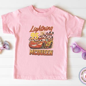May include: Pink short-sleeved t-shirt featuring a graphic of Lightning McQueen from the Cars movie. The design includes the text "Lightning 95 McQueen" in a vintage style, with a checkered flag and the number 95.