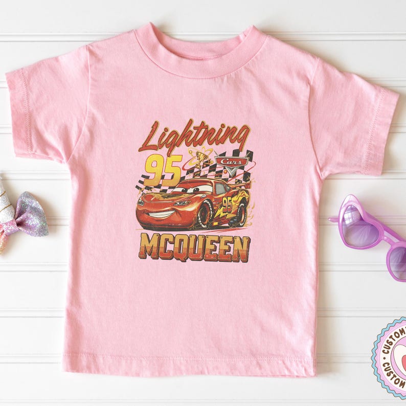 May include: Pink short-sleeved t-shirt featuring a graphic of Lightning McQueen from the Cars movie. The design includes the text "Lightning 95 McQueen" in a vintage style, with a checkered flag and the number 95.