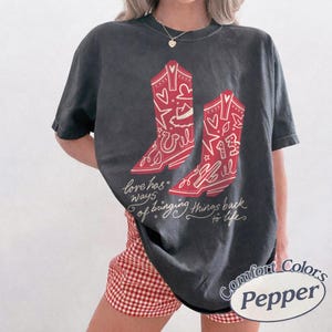 May include: A charcoal grey short-sleeve t-shirt featuring a graphic of two red cowboy boots with white line art details including hearts, stars, and the number 13. The text reads, love has ways of bringing things back to life, in white script.