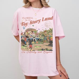 Peut inclure: Un t-shirt rose clair avec un graphisme color&eacute; repr&eacute;sentant une sc&egrave;ne de parc &agrave; th&egrave;me. Le texte indique "Florida Toy Story Land Est. 2018" et "To Infinity and Beyond". Le design comprend des personnages de dessins anim&eacute;s.