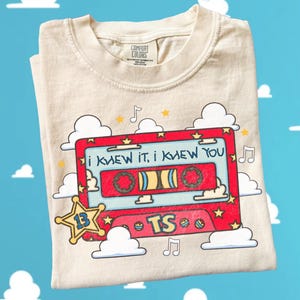 May include: A cream-colored t-shirt featuring a graphic of a red cassette tape with the text "i knew it, i knew you" and "TS". The design includes yellow stars, musical notes, and white clouds, with a yellow star badge containing the number 13.