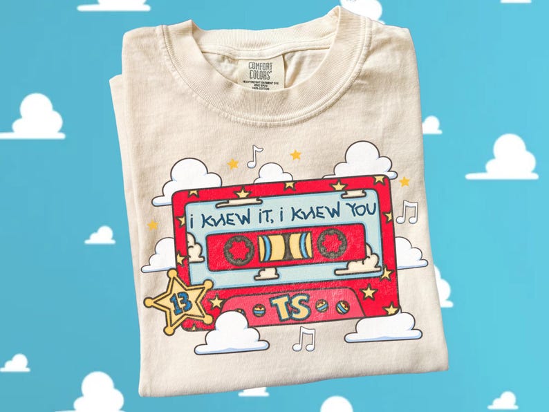 May include: A cream-colored t-shirt featuring a graphic of a red cassette tape with the text "i knew it, i knew you" and "TS". The design includes yellow stars, musical notes, and white clouds, with a yellow star badge containing the number 13.
