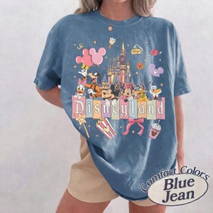 May include: Blue jean-colored t-shirt featuring a colorful Disneyland graphic. The design includes Mickey Mouse, Minnie Mouse, Donald Duck, and other Disney characters, along with the Disneyland castle and the word "Disneyland".