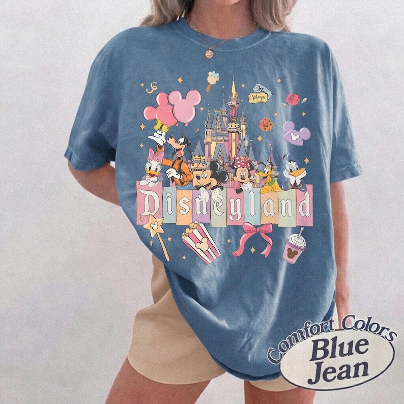 May include: Blue jean-colored t-shirt featuring a colorful Disneyland graphic. The design includes Mickey Mouse, Minnie Mouse, Donald Duck, and other Disney characters, along with the Disneyland castle and the word "Disneyland".