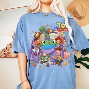 May include: A blue short-sleeve t-shirt featuring a graphic print of Toy Story 5 characters including Woody, Buzz Lightyear, Rex, and Mr. Potato Head emerging from a purple backpack, surrounded by colorful stars and blocks on the front.