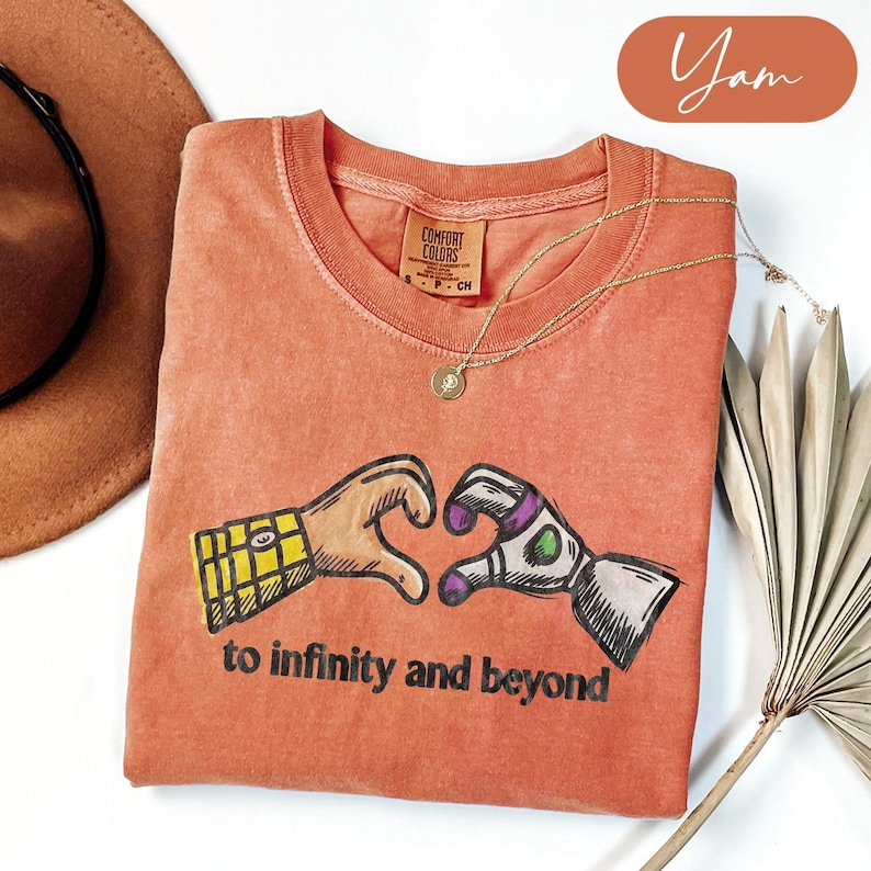 May include: A terracotta-colored crew neck t-shirt featuring a graphic of two hands forming a heart shape, one resembling a cowboy sleeve and the other a space ranger suit. The text "to infinity and beyond" is printed in black below the graphic.