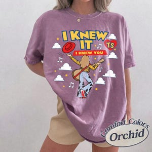 May include: A mauve short-sleeve t-shirt featuring a graphic of a person playing a guitar, wearing a yellow plaid shirt and cow-print trousers. The text reads I KNEW IT I KNEW YOU TS, accompanied by a red hat, a red heart, white clouds, and yellow stars.