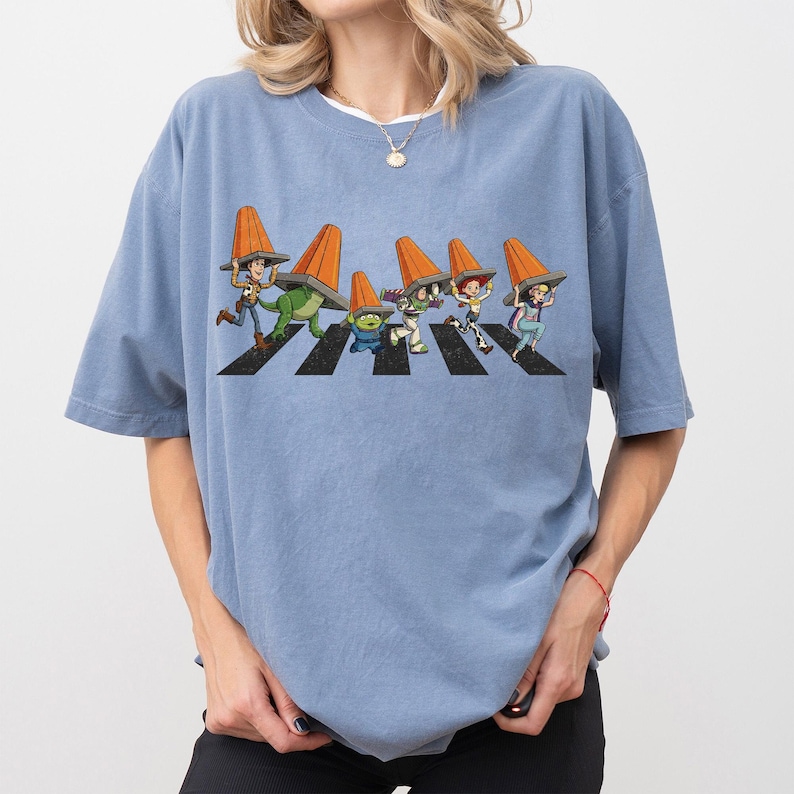 Puede incluir: Camiseta azul claro con un gr&aacute;fico de personajes de dibujos animados cruzando un paso de cebra, cada uno con un cono de tr&aacute;fico naranja. El dise&ntilde;o es una parodia de una famosa portada de &aacute;lbum. La camiseta es holgada.