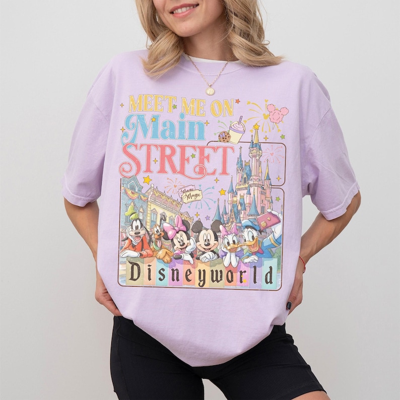 May include: A light purple t-shirt featuring a colorful graphic with the text "MEET ME ON Main STREET Disneyworld". The graphic includes cartoon characters and a castle. The shirt is a casual wear item.