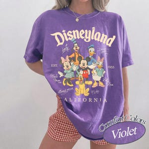 K&ouml;nnte beinhalten: Ein violettes Kurzarm-T-Shirt mit einem Grafikdruck von Disneyland-Figuren, darunter Mickey Maus, Minnie Maus, Donald Duck, Daisy Duck, Goofy und Pluto. Der Text lautet Disneyland, Est. 1955, California sowie die Namen der Figuren.