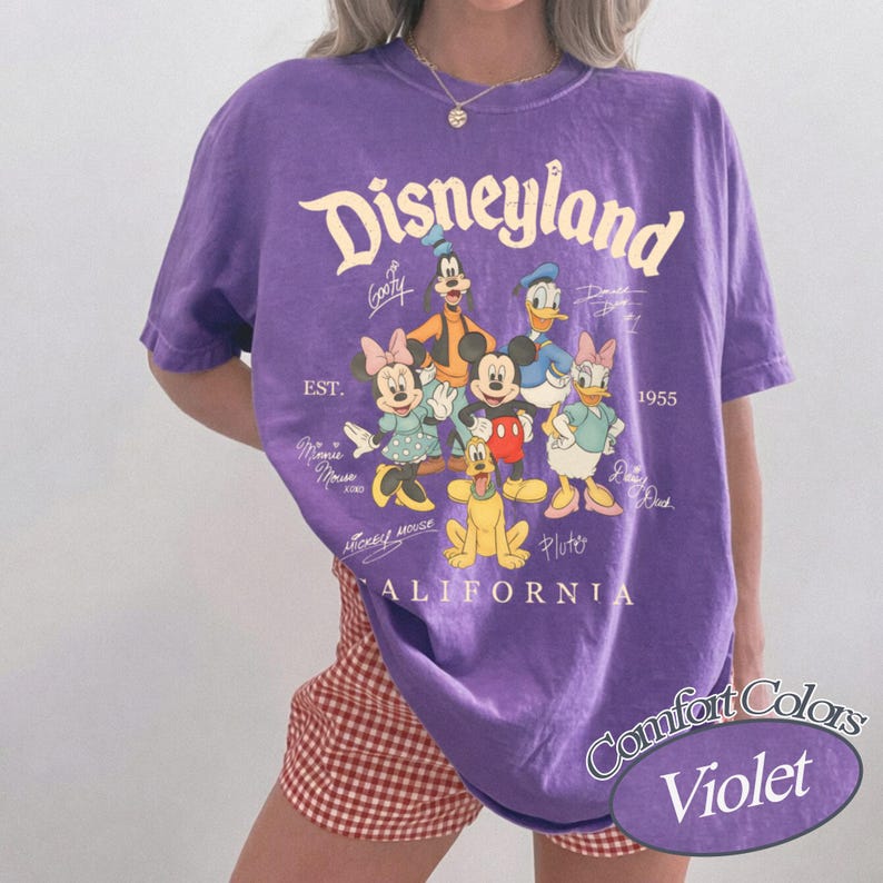K&ouml;nnte beinhalten: Ein violettes Kurzarm-T-Shirt mit einem Grafikdruck von Disneyland-Figuren, darunter Mickey Maus, Minnie Maus, Donald Duck, Daisy Duck, Goofy und Pluto. Der Text lautet Disneyland, Est. 1955, California sowie die Namen der Figuren.