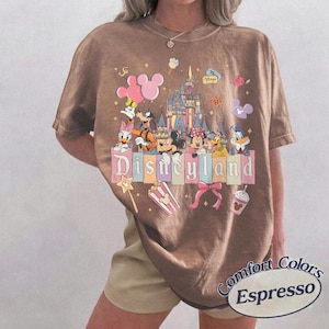 May include: Espresso-colored t-shirt featuring a Disneyland graphic with cartoon characters and a castle. The design includes Mickey and Minnie Mouse, Donald Duck, and Goofy. The text "Disneyland" is displayed prominently. The shirt is from Comfort Colors.