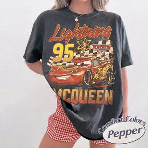 May include: Dark gray t-shirt featuring a graphic of Lightning McQueen from the Cars movie. The design includes the text "Lightning 95 McQueen" in red and yellow. The shirt is a Comfort Colors brand.