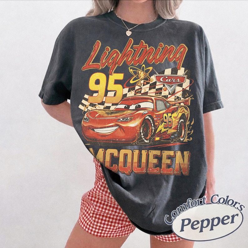 May include: Dark gray t-shirt featuring a graphic of Lightning McQueen from the Cars movie. The design includes the text "Lightning 95 McQueen" in red and yellow. The shirt is a Comfort Colors brand.