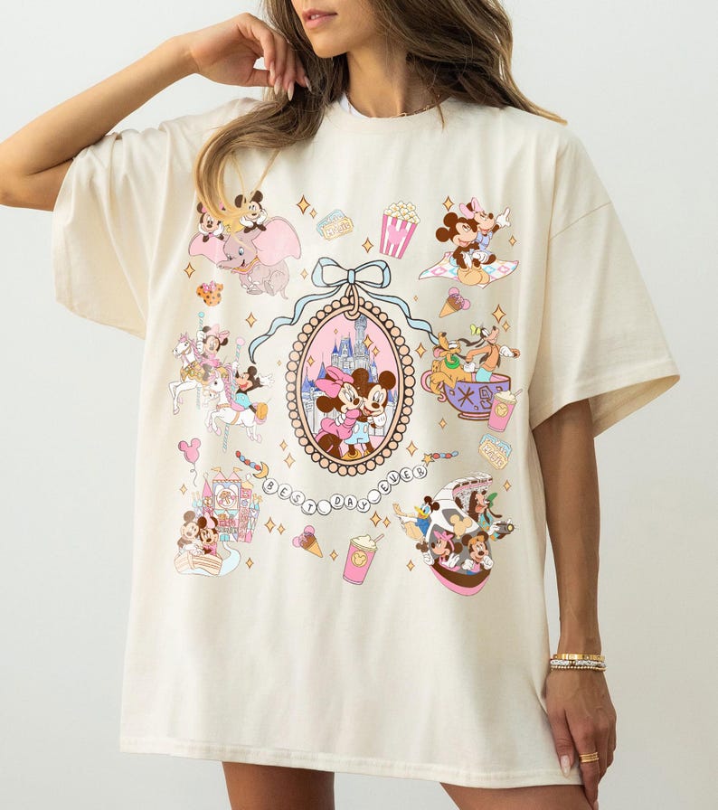 May include: Cream-colored t-shirt featuring a colorful Disney-themed graphic. The design includes illustrations of Mickey and Minnie Mouse, Dumbo, and other characters, along with the text "Best Day Ever."