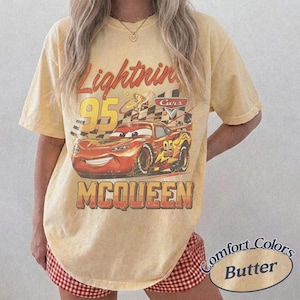 May include: Yellow t-shirt featuring a graphic of Lightning McQueen from the Cars movie. The design includes the number 95, a checkered flag, and the name "McQueen". The shirt is labeled "Comfort Colors Butter".
