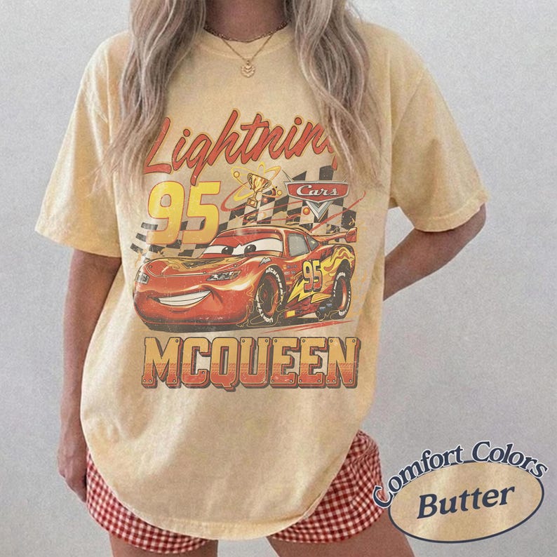 May include: Yellow t-shirt featuring a graphic of Lightning McQueen from the Cars movie. The design includes the number 95, a checkered flag, and the name "McQueen". The shirt is labeled "Comfort Colors Butter".