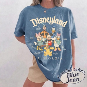 K&ouml;nnte beinhalten: Ein blaues Kurzarm-T-Shirt mit einem Grafikdruck von Disney-Figuren, darunter Mickey Maus, Minnie Maus, Donald Duck, Daisy Duck, Goofy und Pluto. Der Text lautet Disneyland, Est. 1955, California, und enth&auml;lt die Unterschriften der Figuren.