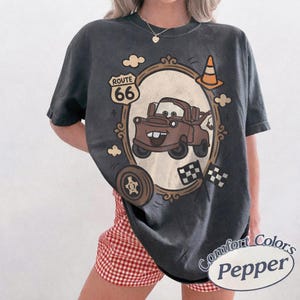 May include: Dark grey t-shirt featuring a cartoon image of Mater from the movie Cars, inside a decorative frame. The shirt includes the text "Route 66" and "Comfort Colors Pepper". Additional graphics include a tire, clouds, and checkered flags.