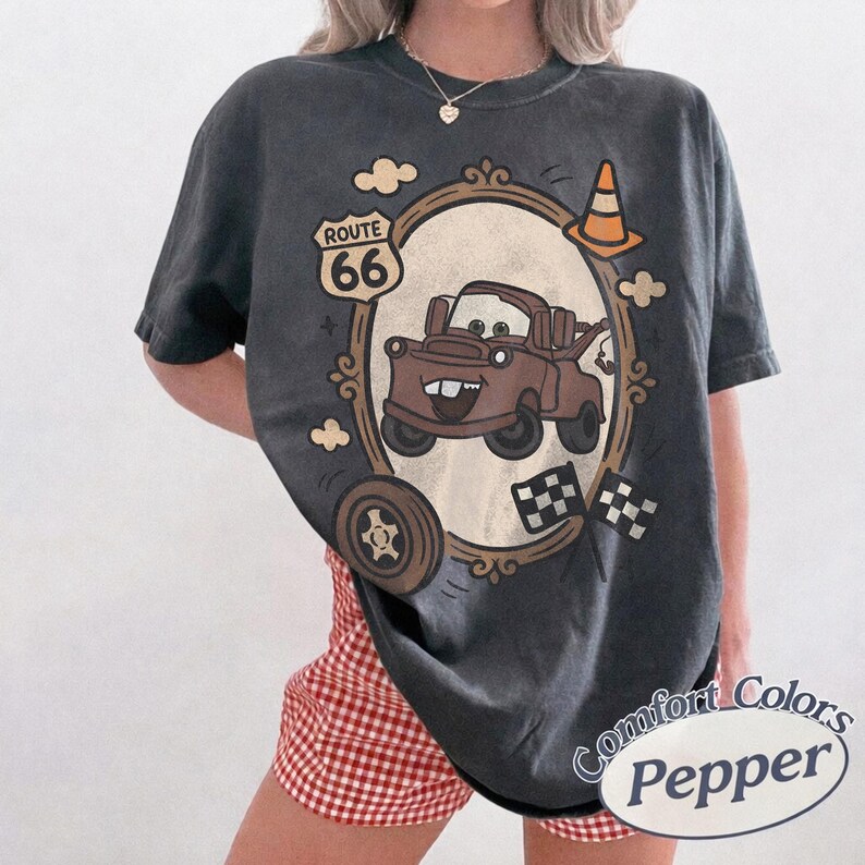 May include: Dark grey t-shirt featuring a cartoon image of Mater from the movie Cars, inside a decorative frame. The shirt includes the text "Route 66" and "Comfort Colors Pepper". Additional graphics include a tire, clouds, and checkered flags.