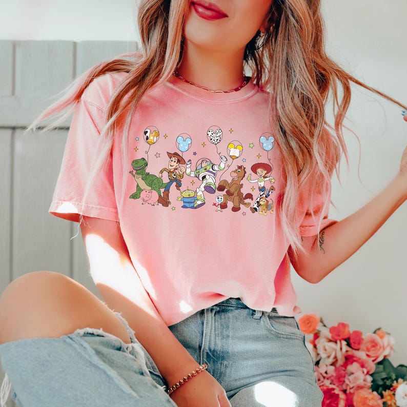 May include: A light pink t-shirt featuring a colorful graphic of animated characters holding balloons. The design includes a green dinosaur, a cowboy, a space ranger, a horse, and a cowgirl. The balloons have star and Mickey Mouse head shapes.