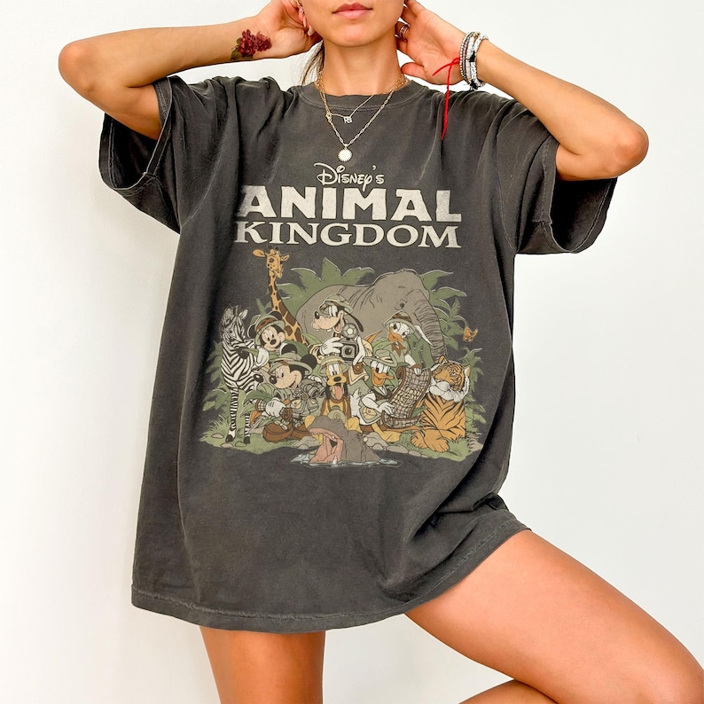 Puede incluir: Camiseta gris oscuro con el texto "Disney's Animal Kingdom" sobre un gr&aacute;fico colorido de varios animales de dibujos animados. El gr&aacute;fico incluye una cebra, una jirafa, un elefante, un tigre y varios personajes de Disney. La camiseta tiene mangas cortas y un corte holgado.
