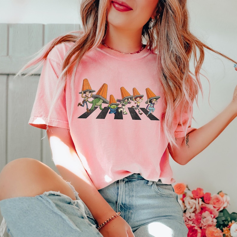 Puede incluir: Camiseta rosa claro con un gr&aacute;fico de personajes de dibujos animados cruzando un paso de cebra, cada uno con un sombrero de cono naranja. El dise&ntilde;o es una parodia de la ic&oacute;nica portada del &aacute;lbum Abbey Road. La camiseta es de material suave.