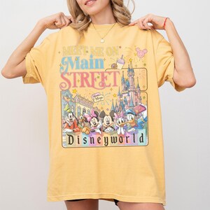 May include: Yellow t-shirt with a colorful graphic featuring Disney characters and the text "Meet Me On Main Street Disneyworld." The design includes a castle, fireworks, and various Disney characters. The shirt is a relaxed fit.