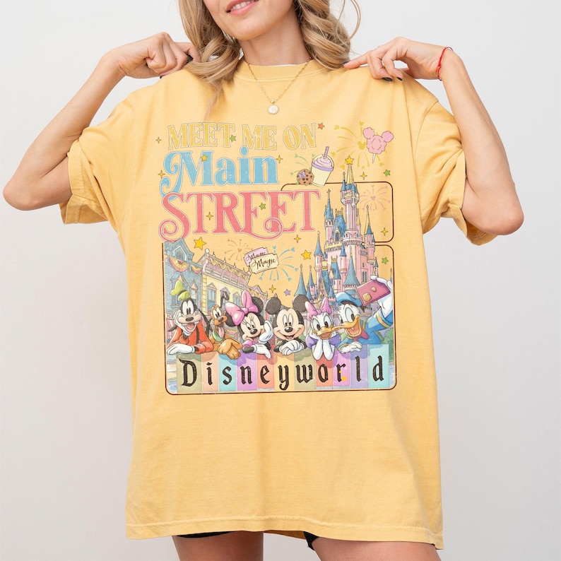 May include: Yellow t-shirt with a colorful graphic featuring Disney characters and the text "Meet Me On Main Street Disneyworld." The design includes a castle, fireworks, and various Disney characters. The shirt is a relaxed fit.