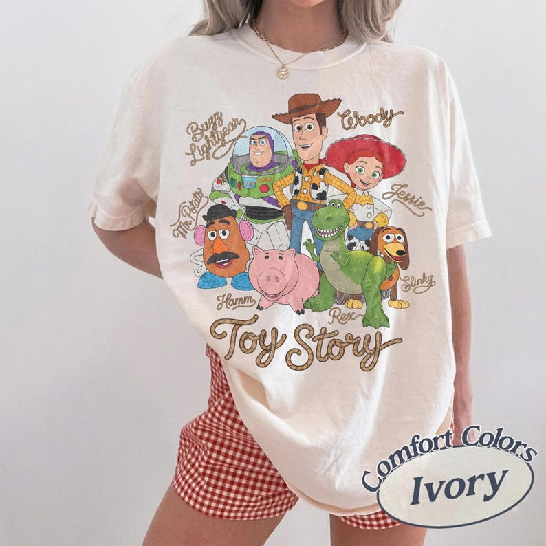 May include: An ivory short-sleeve t-shirt featuring a graphic print of Toy Story characters including Buzz Lightyear, Woody, Jessie, Mr. Potato Head, Hamm, Rex, and Slinky. The text Toy Story is printed below the characters in a brown script font.