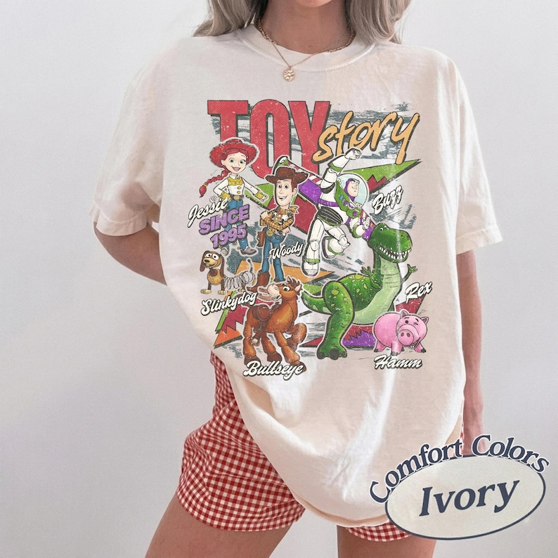 May include: An ivory short-sleeve t-shirt featuring a colorful graphic print of Toy Story characters including Jessie, Woody, Buzz Lightyear, Slinky Dog, Bullseye, Rex, and Hamm. The text reads Toy Story, Since 1995, Jessie, Woody, Buzz, Slinky Dog, Bullseye, Rex, and Hamm.