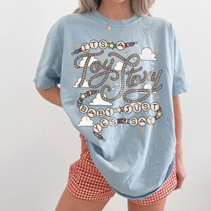 May include: A light blue short-sleeve t-shirt featuring a graphic print with the text "ITS A Toy Story BABY JUST YES SAY". The design includes rope-style lettering, colorful beads, small stars, musical notes, and white cloud shapes.