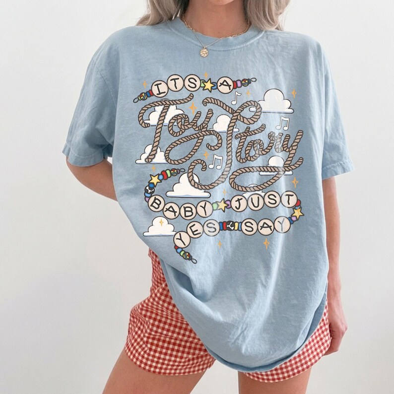 May include: A light blue short-sleeve t-shirt featuring a graphic print with the text "ITS A Toy Story BABY JUST YES SAY". The design includes rope-style lettering, colorful beads, small stars, musical notes, and white cloud shapes.