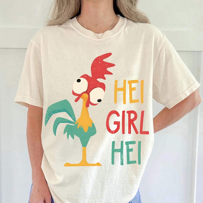 Peut inclure: T-shirt cr&egrave;me avec un coq de dessin anim&eacute; aux accents rouges, jaunes et turquoise. Le texte "HEI GIRL HEI" est imprim&eacute; en jaune, rouge et turquoise. Le t-shirt a une coupe d&eacute;contract&eacute;e et des manches courtes.