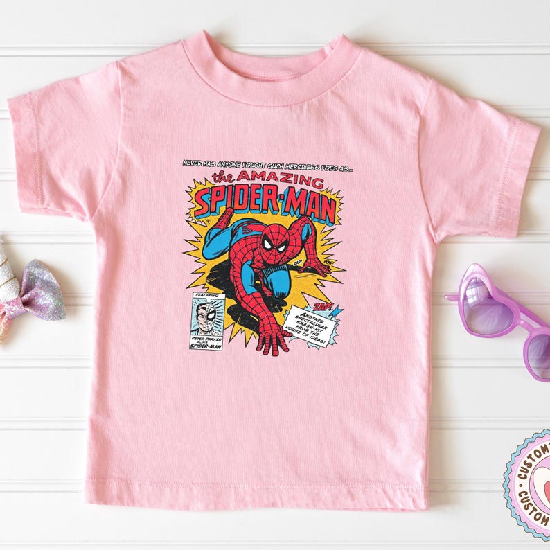 以下が含まれることがあります： スパイダーマンのコミックブックグラフィックが特徴の薄いピンク色のTシャツ。デザインには「The Amazing Spider-Man」の文字やその他のコミック要素が含まれています。このシャツは、クラシックなクルーネックです。