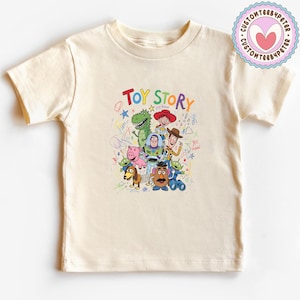 May include: A cream-colored t-shirt featuring a colorful graphic of characters from the animated film "Toy Story." The text "TOY STORY" is written above the characters. The shirt has short sleeves and a crew neck.